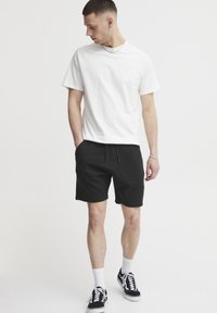 White cotton t-shirt with raglan sleeves, paired with textured black shorts and black sneakers. Model stands sideways with relaxed posture.