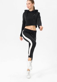 Felpa nera corta con cappuccio e lacci, abbinata a pantaloni jogger neri con una striscia bianca laterale. Sneaker bianche completano il look.