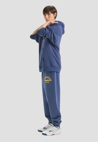 Felpa blu navy con tasca frontale e cordino, abbinata a pantaloni tuta coordinati con un logo giallo. Le sneakers sono bianche con dettagli.