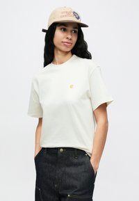 Ženska z dolgimi črnimi lasmi, ki nosi bež kapa, belo majico s kratkimi rokavi z majhnim logotipom in temne kavbojke z rokami v žepih.