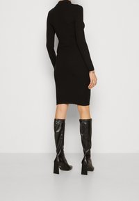 Robe noire à manches longues côtelées, cintrée à la taille avec une longueur genoux, associée à des bottes brillantes noires à talons arrivant jusqu'aux genoux.