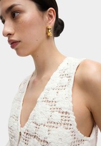 Goldene skulpturale Ohrringe, strukturierter floraler Spitze-Oberteil in Weiß mit tiefem V-Ausschnitt und Knopfdetails, die Haut unter dem Design zeigen.