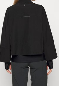 Chaqueta negra corta con mangas largas, que presenta un cuello alto, una abertura en la parte trasera y acentos reflectantes. Textura suave y diseño minimalista.