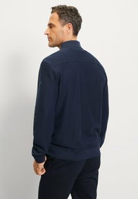 Marineblaue Fleecejacke mit hohem Kragen, langen Ärmeln und einer sanften Textur. Verfügt über ein kleines Logo am linken Arm.