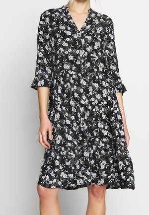 Kvinde iført sort knælængde kjole med hvidt blomsterprint, trekvartærmer og knapper ned foran på livdelen.