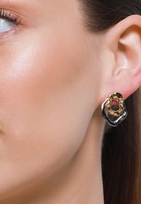 Orecchini astratti in oro e argento con una forma ondulata e texture. Il design presenta un ritaglio al centro che mette in risalto dettagli intricati.