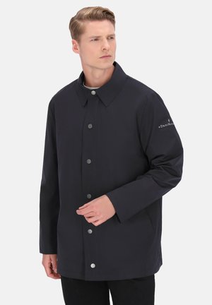 Sötétkék, vízálló dzseki egyenes szabással, nagy gallérral, elülső patentgombokkal és logóval a bal ujján. Sima textúra.