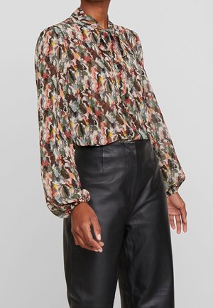 Femme portant un chemisier à motif abstrait multicolore avec un col noué et des manches longues bouffantes, associé à un pantalon en cuir noir taille haute.