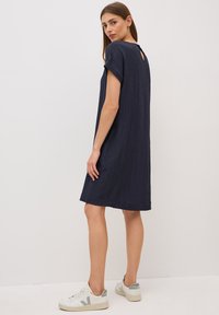 Mörkblå knälång klänning i en texturerad tyg, korta ärmar och en nyckelhålsdetalj på baksidan, kombinerad med vita sneakers.