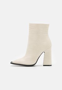 Białe botki sięgające kostki z szpiczastym noskiem, teksturowanym wzorem krokodyla i blokowym obcasem. Podeszwa ma czarny akcent z przodu.