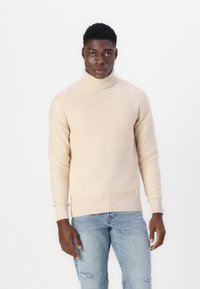 Maglione beige con collo a dolce vita e polsini e orlo a costine, realizzato in un morbido tessuto a maglia. Indossato con jeans chiari strappati.