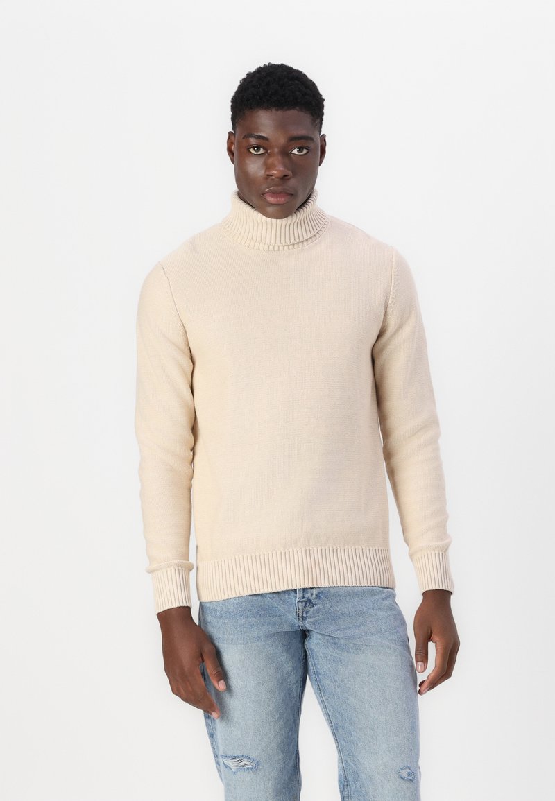 Maglione beige con collo a dolce vita e polsini e orlo a costine, realizzato in un morbido tessuto a maglia. Indossato con jeans chiari strappati.