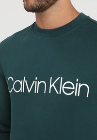 Muž v tmavě zelené mikině s kulatým výstřihem, na které je velké bílé logo "Calvin Klein" přes hrudník, zobrazuje dolní část obličeje a krk.