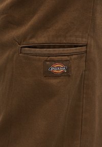 Tissu marron avec une petite poche passepoilée ornée d'une étiquette rectangulaire cousue de la marque Dickies, aux couleurs marron, rouge, jaune et bleu.