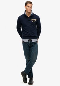 Suéter azul marino con cremallera, logo "SUPERDRY N.Y.C.", superpuesto sobre una camisa de cuadros clara. Combinado con jeans azules y zapatos de cuero negros.