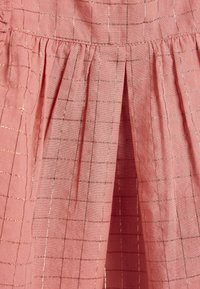 Tissu rose avec de fins fils d'or horizontaux et verticaux formant un motif en grille, avec des plis cousus près du bord supérieur.