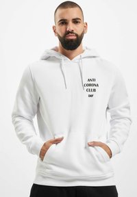 Weißer Hoodie aus weichem Material, mit einer frontalen Kängurutasche und schwarzem Text "ANTI CORONA CLUB DEF", der zentral auf der Brust aufgedruckt ist.