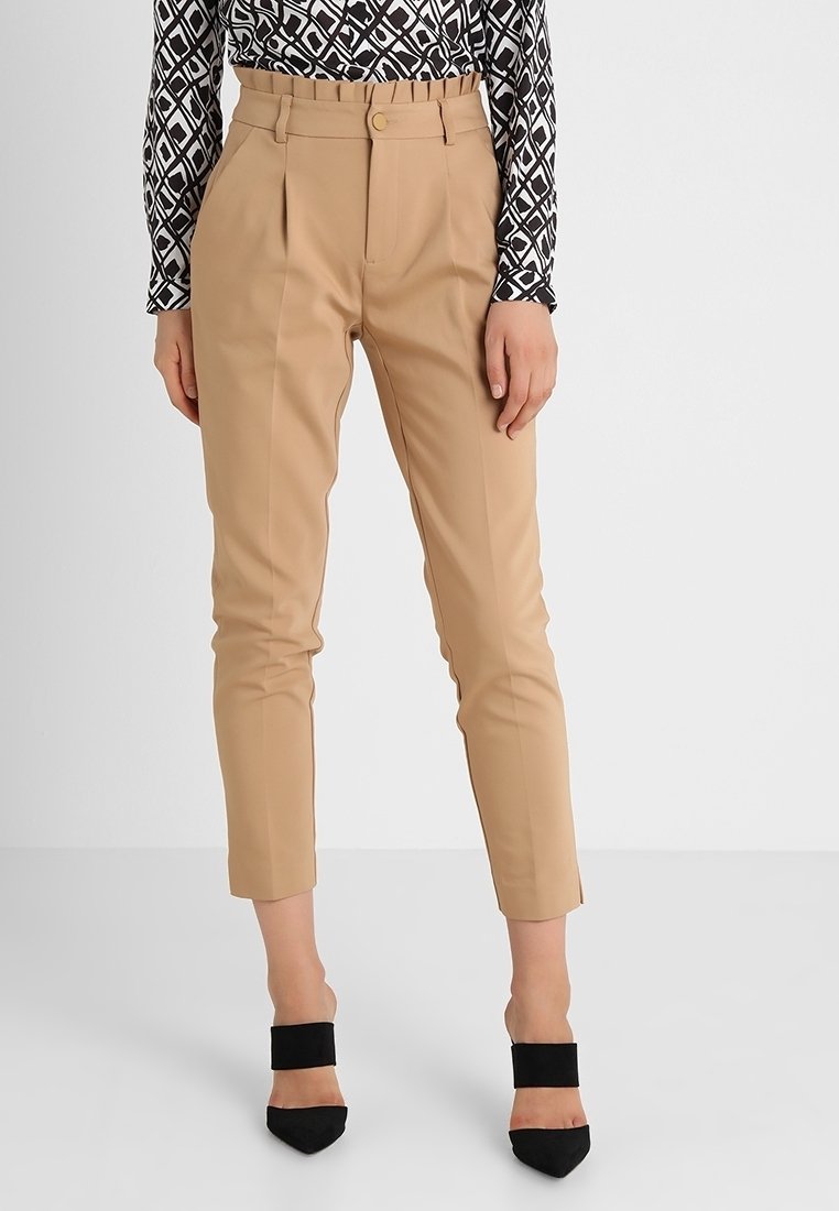 Beige højtaljede tapered bukser med frontfolder, båret med sorte spidse hæle og en langærmet top med sort-hvide mønstre.