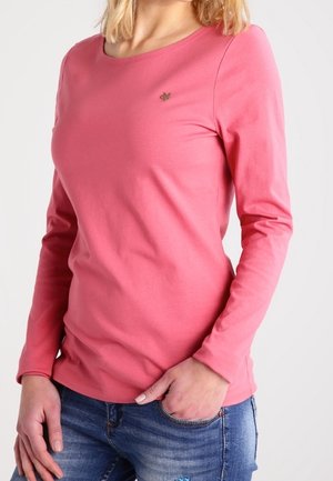 Vrouw draagt een nauwsluitend roze shirt met lange mouwen en een klein geborduurd logo op de borst en blauwe versleten jeans.