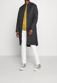 Abrigo acolchado negro con cuello redondo, botones a presión, llevado sobre un suéter mostaza y pantalones blancos, combinado con zapatillas verde oliva.