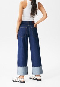 Jeans de denim de pierna ancha con un lavado azul oscuro, dobladillo en denim más claro y dos bolsillos traseros. Llevados con sandalias de tiras en blanco y negro.