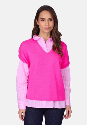 Femme portant un pull sans manches rose vif à col en V, sur une chemise rayée rose clair à boutons, avec un pantalon foncé, debout devant un fond uni.