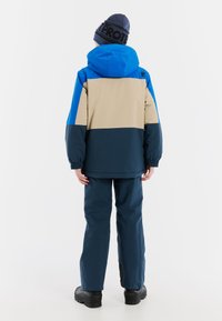 Kolorowa zimowa kurtka w niebiesko-beżowo-granatowym wzorze z blokami kolorów, z elastycznymi mankietami i dużym kapturem; zestawiona z ciemnoniebieskimi spodniami.