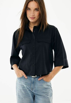 Donna con capelli lunghi castani che indossa una camicia nera con bottoni e maniche arrotolate e jeans azzurri, con le mani in tasca, su sfondo bianco.