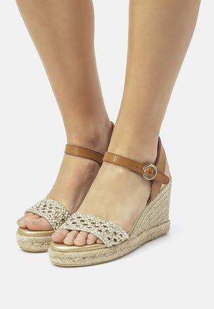 Keilsandalen mit einem braunen Knöchelriemen, gewobenem beigen Obermaterial und einer strukturierten Espadrille-Sohle. Das Design verfügt über offene Zehen und ein dekoratives Muster.