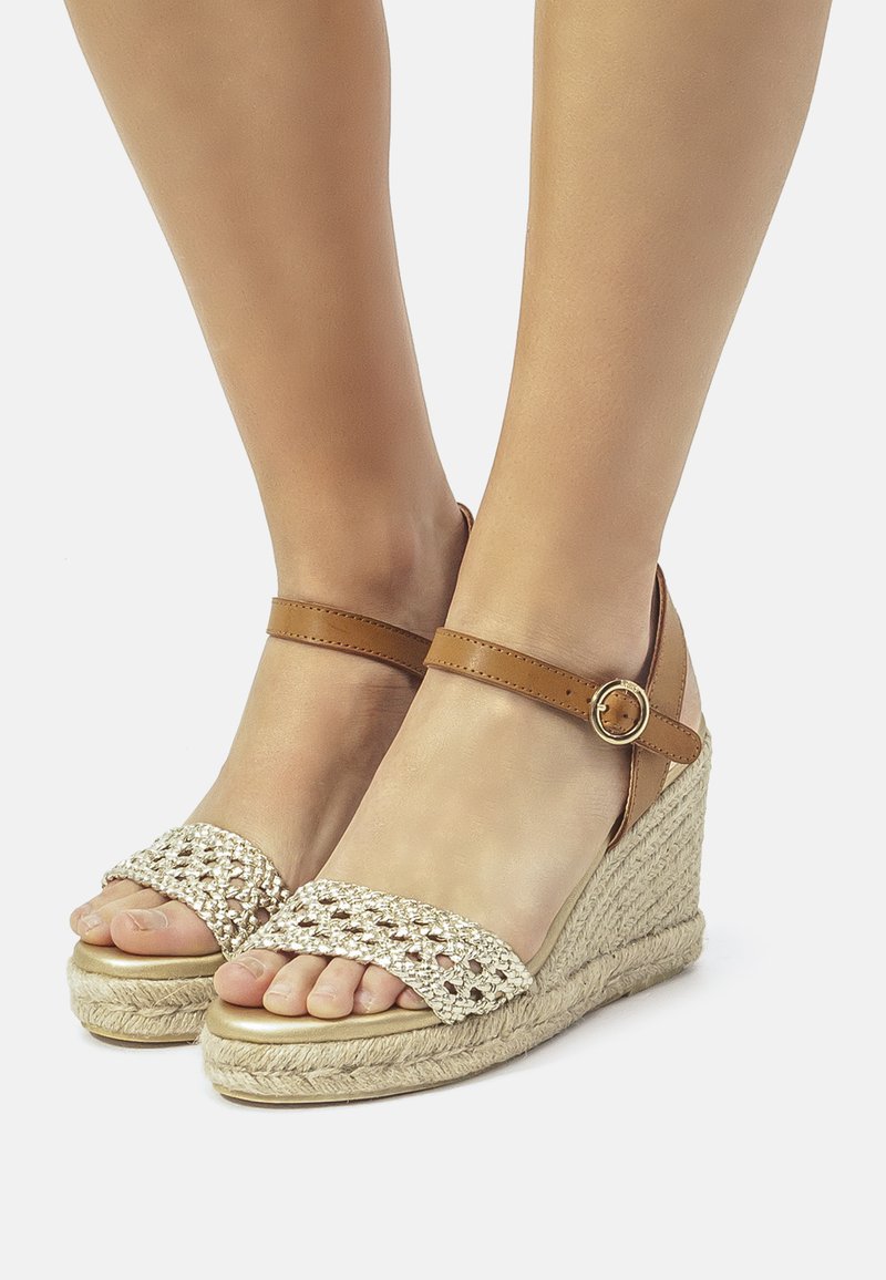 Keilsandalen mit einem braunen Knöchelriemen, gewobenem beigen Obermaterial und einer strukturierten Espadrille-Sohle. Das Design verfügt über offene Zehen und ein dekoratives Muster.