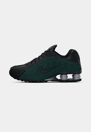Zapatilla deportiva en negro y verde oscuro con parte superior de malla y sintético, diseño curvado distintivo y amortiguación de aire visible en la suela.