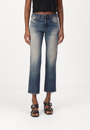 Frau trägt ausgewaschene, gerade geschnittene blaue Jeans, kombiniert mit schwarzen offenen Schuhen mit goldenen Schnallen, stehend vor einem einfarbigen Hintergrund.