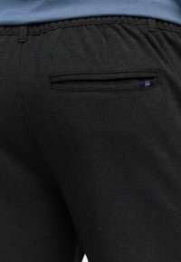 Nahaufnahme von schwarzen Hosen mit elastischem Bund, einer hinteren Paspeltasche und einem kleinen Stoffetikett mit weißem Logo auf Marineblau.