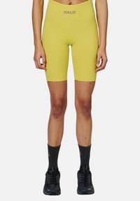 Geel fietsshorts met een hoge tailleband en het "HALO" logo. Draagt met zwarte sportschoenen en donkere sokken met gele tekst.