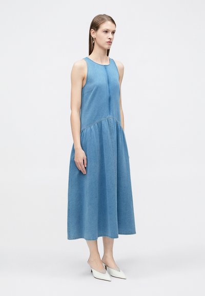 Γυναίκα που στέκεται, φοράει αμάνικο μπλε μίντι φόρεμα από τζιν ύφασμα και λευκά, μυτερά παπούτσια με χαμηλό τακούνι, σε απλό φόντο.