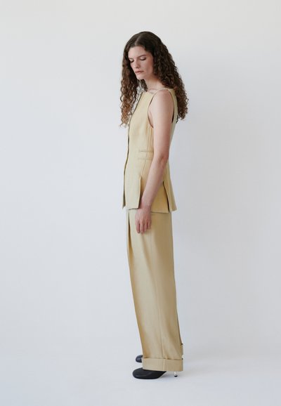 Γυναίκα με σγουρά μαλλιά που φοράει μπεζ αμάνικο γιλέκο και παντελόνια με φαρδιά πόδια ταιριαστά, στέκεται πλευρικά μπροστά σε ένα απλό φωτεινό φόντο.