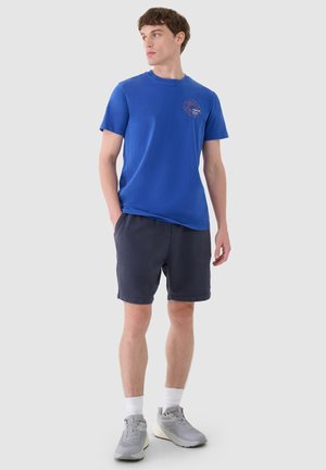 Mężczyzna stoi z rękami w kieszeniach, ubrany w niebieski T-shirt z nadrukiem, ciemne szorty, białe skarpetki i szare tenisówki, patrzy na bok.