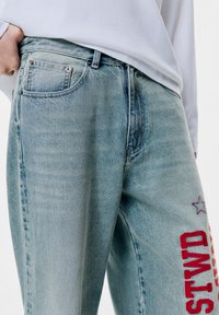 Ljusa blå jeans i denim med avslappnad passform, prydda med ett rött broderat motiv och stjärndetaljer. Inkluderar fickor fram och bak.