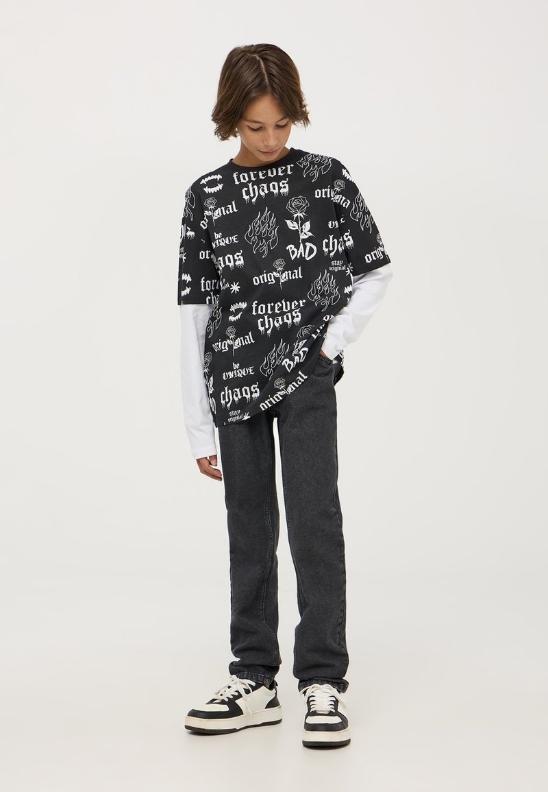 T-shirt grafica nera con testo e disegni bianchi, indossata sopra una maglietta a maniche lunghe bianca. Abbinata a jeans in denim scuro e sneakers nere.