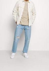 Giacca di jeans bianca sopra una maglietta beige, abbinata a jeans azzurri chiari con strappi. Sneakers bianche completano l'outfit.