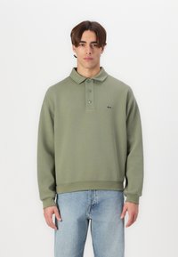 Sudadera verde oliva con cuello tipo polo y cierre de botones, que presenta puños y bajo acanalados, y un pequeño logo en el lado izquierdo del pecho.