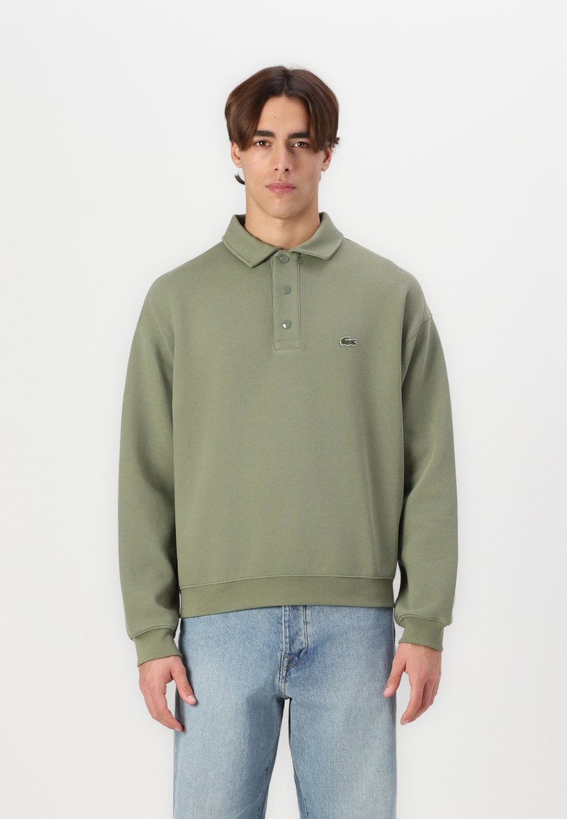 Sudadera verde oliva con cuello tipo polo y cierre de botones, que presenta puños y bajo acanalados, y un pequeño logo en el lado izquierdo del pecho.