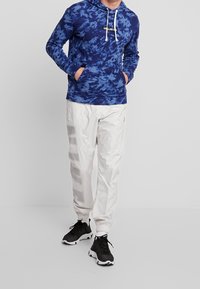 Hoodie bleu tie-dye avec cordons blancs et logo sur le devant, associé à un pantalon blanc comportant un grand imprimé gris et des poignets élastiques, porté avec des baskets noires.