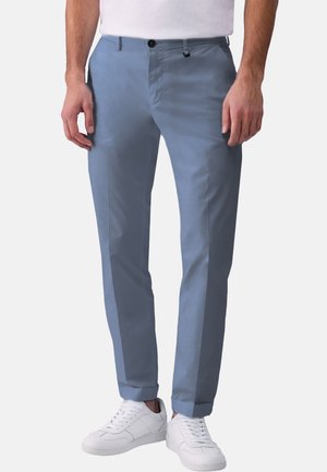 Uomo che indossa pantaloni slim fit azzurri chiari con orli risvoltati e sneakers bianche, in piedi davanti a uno sfondo semplice.