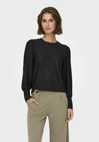 Top nero a maniche lunghe con un motivo testurizzato e scollatura rotonda, abbinato a pantaloni khaki con tasche laterali e dettaglio a bottone.