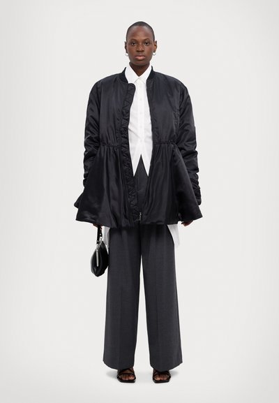 Schwarze Satinstaffeljacke mit gerafftem Bund, weißes Hemd mit Knöpfen, graue weite Hose und schwarze Handtasche. Beinhaltet flache Sandalen.