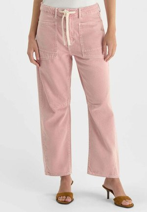 Vrouw draagt een hoge taille, lichtroze wijdbeeldige broek met grote voorzakken en beige trekkoord, gecombineerd met bruine open-toe sandalen met hak.