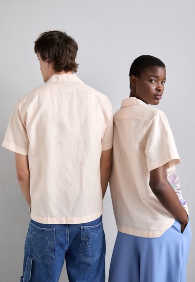 Short-sleeve, light peach shirts made of textured fabric, featuring a collar, worn by two models, with one displaying a colorful graphic.