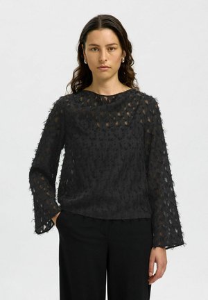 Blouse noire texturée à coupe décontractée, avec des manches longues, un col bateau, et un design superposé et transparent avec des bords effilochés.