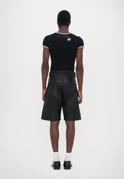 Schwarzes Kurzarmhemd mit lila Akzenten und Text auf der Rückseite. Schwarze Ledershorts mit einem Gürtel und Gesäßtaschen. Schwarze Schuhe und weiße Socken.